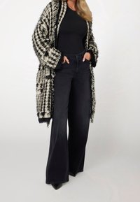 Una donna vestita con jeans neri a gamba larga, una maglietta nera e un cardigan frangiato a scacchi bianchi e neri, in piedi con le mani nelle tasche.