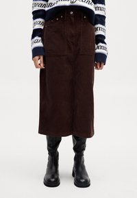 Gonna midi in corduroy marrone con due tasche anteriori, abbinata a stivali neri fino al ginocchio e a un maglione a strisce. La gonna ha un taglio dritto.