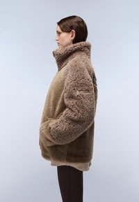 Lažno krzno oversized jakna v svetlo rjavi barvi z visokim ovratnikom, stranskimi žepi in teksturiranimi rokavi. Mehak, puhast material z gladko površino.