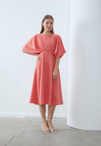 Korallfärgad knälång klänning i lätt material med korta, svävande ärmar och en avslappnad passform. Matchas med strappy vita sandaler.
