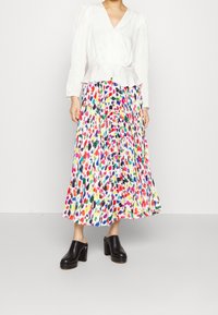 Blusa blanca con cintura peplum, combinada con una falda maxi plisada de varios colores que presenta patrones abstractos llamativos. Zapatos negros de tacón grueso.