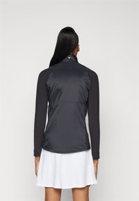 Černá, lehká vesta s vysokým límcem, s hladkou, lesklou texturou. Dlouhé rukávy pod vestou a přiléhavý design.