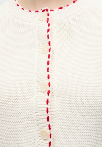 Gros plan d'un cardigan blanc en maille avec des boutons crème et une bordure cousue rouge le long du col et de la ligne de boutons.