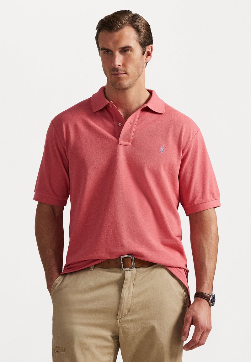 Κοράλι πόλο μπλούζα με κοντά μανίκια και γιακά, που διαθέτει ένα μικρό μπλε έμβλημα. Φορεμένο με μπεζ παντελόνι και καφέ ζώνη, παρουσιάζοντας μια άνετη εμφάνιση.