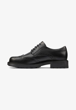 Chaussure brogue en cuir noir avec perforations en forme d’aile, fermeture à lacets et semelle épaisse crantée, vue de profil sur fond blanc.