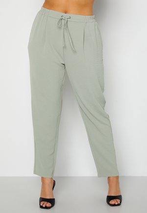 Mujer con pantalones verdes claros con cordón y tacones negros con dedos abiertos, de pie contra un fondo blanco liso.