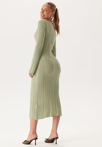 Robe maxi à manches longues en tissu vert doux, avec une silhouette ajustée et une fente sur le côté. Associée à des mules à talons à imprimé léopard noir.