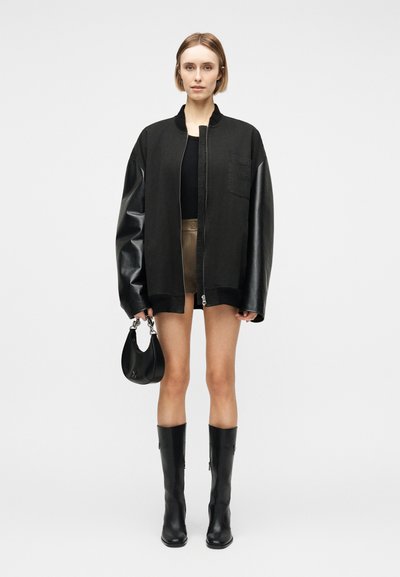 Femme aux cheveux courts portant une veste noire oversize avec des manches en cuir, un short beige, un haut noir, des bottes noires, tenant un petit sac à main noir.