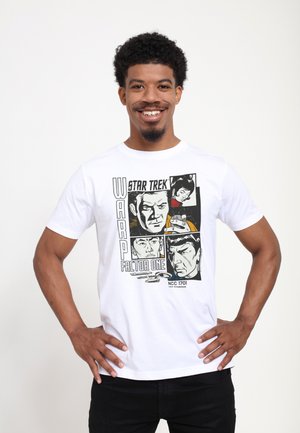 Lächelnder Mann trägt ein weißes Star Trek T-Shirt mit schwarz-weißen Comic-Stil Crew-Porträts und dem Text „Warp Factor One“ und steht mit den Händen in den Hüften.