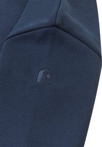 Granatowa tkanina o teksturowej powierzchni z wytłoczonym logo w dolnej części. Materiał jest miękki i dopasowujący się do sylwetki.