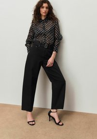 Schwarze durchsichtige Bluse mit Punkten, mit Kragen und Knöpfen, kombiniert mit hoch taillierter schwarzer Hose und schwarzen, mit Riemchen versehenen Absätzen.