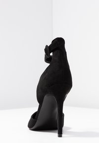 Černá semišová lodička na vysokém podpatku se štíhlým stiletto podpatkem a páskem se zaoblenou sponou kolem kotníku. Hladká textura, elegantní silueta.