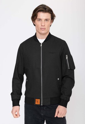 Junger Mann trägt eine schwarze Bomberjacke mit Front-Reißverschluss und Reißverschlusstasche am linken Ärmel und steht vor einem weißen Hintergrund.
