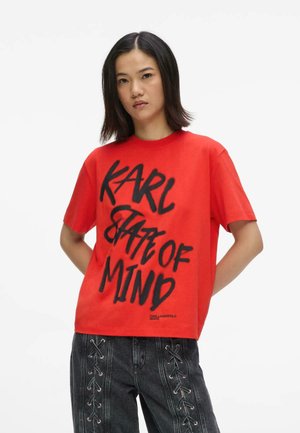 Jeune femme portant un t-shirt rouge avec le texte « KARL STATE OF MIND » et un jean noir avec des détails à lacets à l'avant.