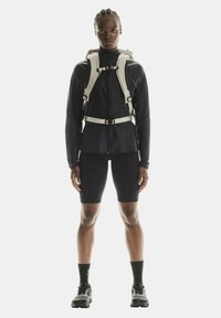 Veste technique noire avec fermeture éclair, sangles blanches ajustables et un sac à dos. Un short noir ajusté et des baskets noires texturées complètent le look.