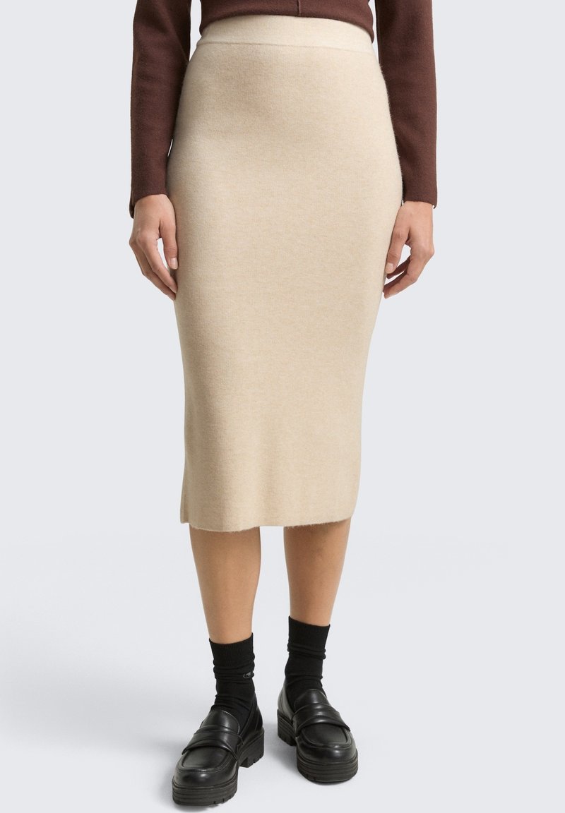 Beżowa dzianinowa spódnica ołówkowa o dopasowanym kroju, o długości do połowy łydki, z prążkowanym paskiem. Noszona z czarnymi skarpetkami i mokasynami na grubej podeszwie.