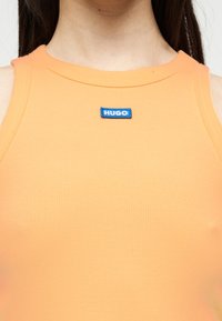 Κοντινό πλάνο ατόμου που φοράει πορτοκαλί αμάνικη ριγέ μπλούζα με μια μικρή μπλε ετικέτα «HUGO» στο λαιμό.