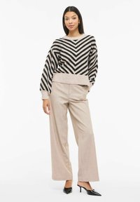 Pull en maille à rayures chevrons noires et crème avec manches larges, associé à un pantalon taille haute beige clair et des escarpins noirs pointus.