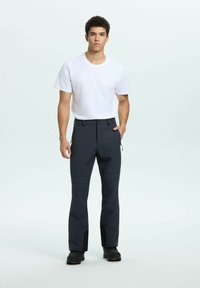 Ung man som står, iklädd en enkel vit t-shirt, mörka träningsbyxor med dragkedjefickor och svarta skor mot en ljus bakgrund.