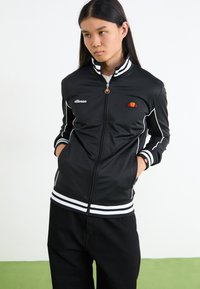 Sort zip-up jakke med hvide striber på kraven og manchetterne, der har et logo-patch på brystet samt sidelommer. Glat, atletisk materiale.