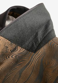 Cuello de una prenda que presenta una sección de fieltro negro texturizado y una tela estampada con tonos tierra, mostrando líneas onduladas en bronce y negro.