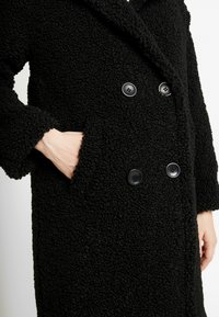 Černý dvouřadý kabát vyrobený z texturované tkaniny. Obsahuje velké černé knoflíky a klopový límec. Viditelné boční kapsy.