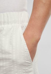 Biely textúrovaný materiál s vertikálnymi pruhmi, s elastickým pásom a detailom bočnej vrecka. Detailný záber na materiál a strih.