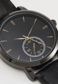 Black analog watch with a round case, gold hour markers, and a subdial. Features a textured black leather strap and a smooth glass face.