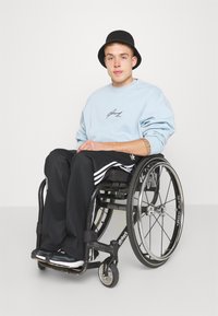 Jeune homme portant un chapeau noir, un sweat-shirt bleu clair et un pantalon noir, assis dans un fauteuil roulant noir sur un fond uni.