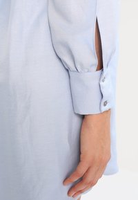 Camicia a maniche lunghe blu chiaro che mostra un tessuto strutturato, polsini aderenti con dettagli di bottoni e aperture laterali per un tocco di stile e comfort aggiuntivo.