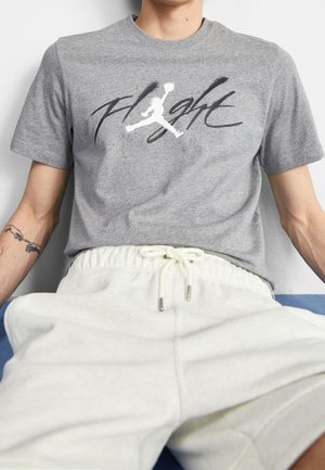 Person, die ein graues T-Shirt mit weißem Jumpman-Basketball-Logo und dem Wort "Flight" in schwarzer Handschrift trägt, kombiniert mit weißen Hose mit Kordelzug.