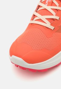 Orange athletic shoe with a textured mesh upper, flat beige laces, and a white and pink rubber sole. Features a sleek design.