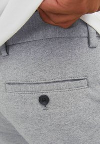 Šedá texturovaná látka kalhot s zadní kapsou se černým knoflíkem a šitými detaily v blízkosti pasového lemu.