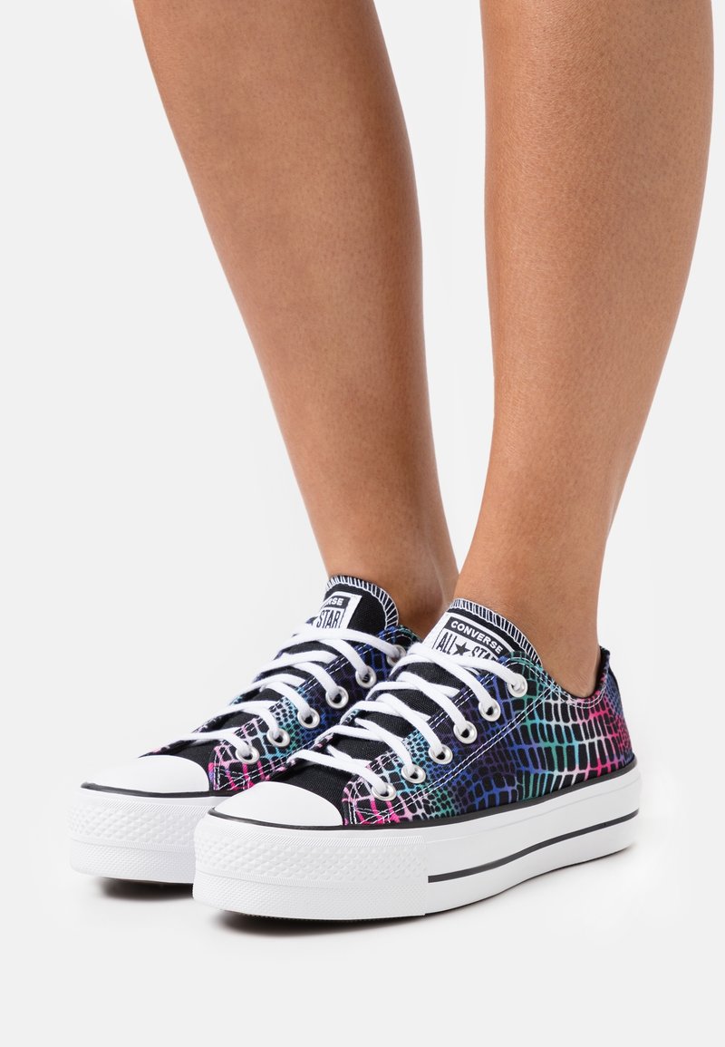 Jambes portant des baskets Converse All Star basses avec un motif d’écailles noir, blanc et multicolore rose, bleu et vert, et des lacets blancs.