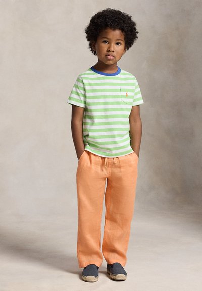 Niño con cabello rizado que lleva una camiseta de rayas verdes y blancas, pantalones naranja y zapatos oscuros sin cordones, de pie con las manos en los bolsillos.