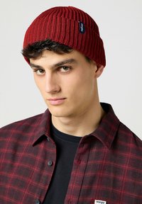 Berretto rosso lavorato a maglia con texture a coste e un piccolo etichetta del marchio. Indossato sopra una maglietta nera e una camicia flanella a quadratini rossi.