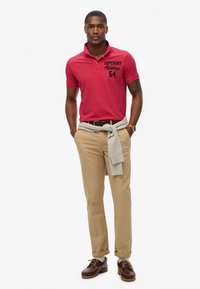 Rød bomulds polo-shirt med sort logo, parret med beige bukser, grå sweater bundet om livet og brune snørebåndssko.