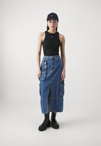 Black tank top, denim cargo skirt with front slits, pockets, and zippers. Black cap with logo. Chunky black boots. Minimalist background.