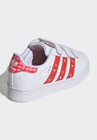 Biele tenisky s tromi červenými pásikmi so srdcovými vzormi, s prednými suchými zipsami a štruktúrovanou gumovou podrážkou. Zlatý logotyp na päte.