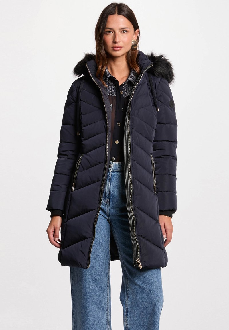 Cappotto imbottito navy in stile trapuntato con cappuccio in pelliccia sintetica, chiusura a zip e tasche laterali. Presenta un taglio lungo e dritto e pannelli texturizzati.