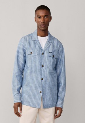 Homme portant une chemise boutonnée à rayures verticales bleues et blanches sur un t-shirt blanc, associée à un pantalon clair, debout devant un fond neutre.