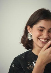 Junge Frau mit kurzen dunklen Haaren lächelt mit geschlossenen Augen, trägt große rechteckige Kristallohrringe und ein schwarzes Oberteil mit Paillettenverzierungen.