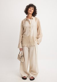 Beige transparante blouse met lange mouwen en knopen, gecombineerd met losse beige broek. Houdt een kleine handtas vast met een contrasterende rand. Sandalen maken de look af.