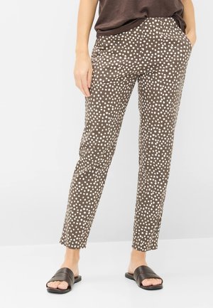 Person iført brune bukser med hvide uregelmæssige polkaprikker, mørkebrun skjorte og sorte slidersandaler, stående med hænderne i lommerne.