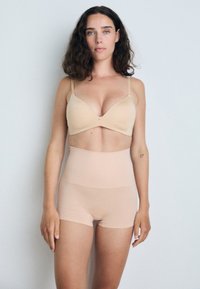 Bralette beige senza cuciture con spalline regolabili e una sottile decorazione centrale, abbinata a pantaloni corti trasparenti beige a vita alta. Texture liscia.