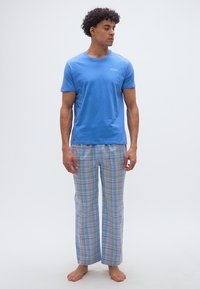 Άνδρας που στέκεται ξυπόλυτος φορώντας μπλε μπλουζάκι πόλο και πιτζάμες με παστέλ καρό, κοιτάζοντας προς τα δεξιά του με λευκό φόντο.