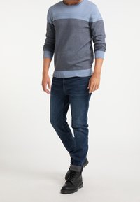 Homme portant un pull bleu clair et gris, un jean bleu foncé retroussé aux chevilles et des bottes noires à lacets sur un fond neutre.