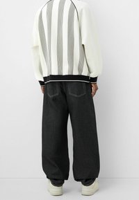 Sudadera blanca con rayas verticales en negro y gris, puños y dobladillo acanalados en negro. Pantalones de mezclilla oscuros y holgados, combinados con zapatillas blancas.