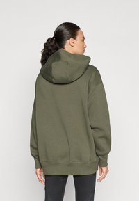 Sudadera verde oliva con una gran capucha, corte holgado, puños y dobladillo acanalados, hecha de una tela suave. Se lleva con leggings negros.