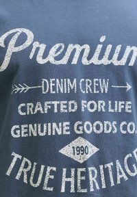 Blått tyg med vit slitna texten: "Premium Denim Crew Skapat för livet Genuine Goods Co. 1990 Äkta arv."
